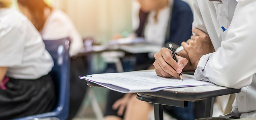 Students seated in a classroom, focused on writing exams. A person in the foreground is holding a pen, writing on paper, with others blurred in the background, indicating an academic setting.