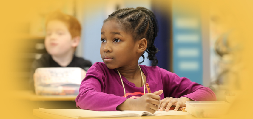 A young girl with braided hair sits at a desk, holding a pencil and looking attentively to her right. She is wearing a purple shirt. In the blurred background, another child is seated at a desk. The image has a soft yellow border.