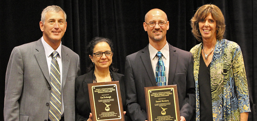 Four people stand together, two of them holding plaques. They are dressed in formal attire, smiling, with a dark backdrop behind them.