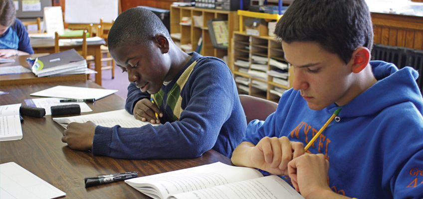Two students sit at a wooden table in a classroom, focused on reading books. One wears a striped sweater and the other a blue hoodie. Various classroom materials are visible in the background.