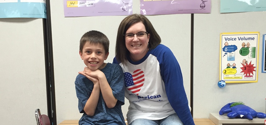 A smiling boy sits on a desk, posing with hands under his chin, next to a smiling woman wearing glasses and a white long-sleeve shirt with a heart-shaped American flag. They are in a classroom with educational posters on the wall.