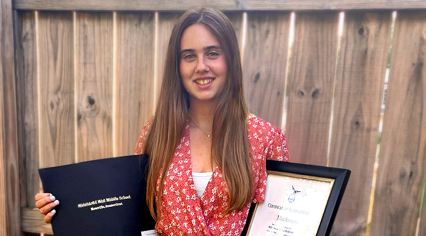 A young woman with long brown hair stands in front of a wooden fence, smiling. She wears a red floral dress and holds a black diploma cover and a framed certificate.