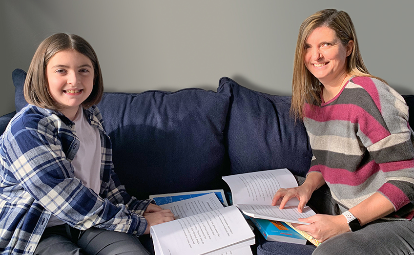 A woman and a young girl sitting on a blue couch, both smiling. The woman is wearing a striped sweater, and the girl is in a checkered shirt over a white tee. They are looking at papers and a book together on the couch.