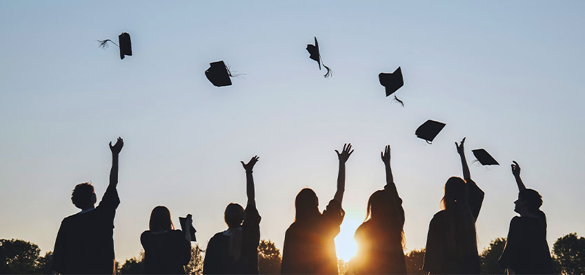 Silhouetted graduates toss their caps into the air against a sunset sky. Seven people stand side by side in celebration, with trees visible in the background.