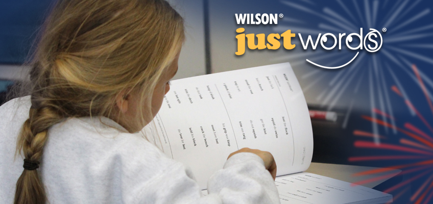 A young girl with light hair is sitting and looking at a workbook. The cover says 