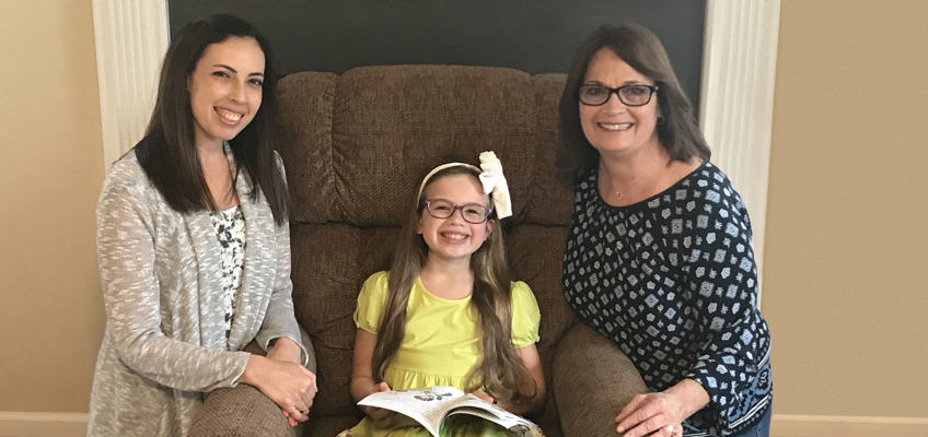 A young girl with glasses and a large bow in her hair sits on a brown armchair holding a book, smiling between two women who are kneeling beside her. Both women are smiling, with one wearing a patterned dress and the other in a cardigan.