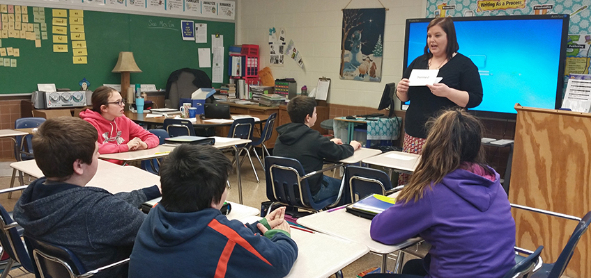 A teacher stands in front of a classroom, holding a flashcard and talking to a group of seated students. The room is filled with desks, educational materials, and a digital screen displaying a blue background.