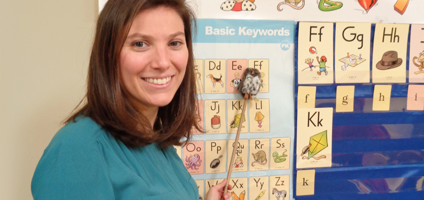 A woman stands smiling next to a colorful educational wall display featuring the alphabet and illustrations. She holds a pointer with a small plush animal attached, pointing at the letter 'J'.
