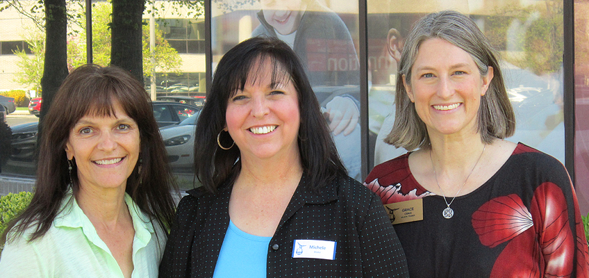 Three women smiling outdoors in front of a window. Two women have dark hair, and the third has gray hair. They're dressed in casual business attire. Trees and parked cars are visible in the background.