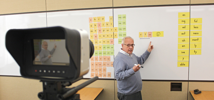 A person is standing in front of a whiteboard with colorful letter and word cards. They point to a section with a marker while being filmed by a video camera in the foreground.