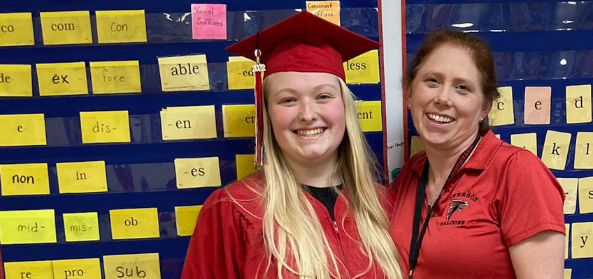 A smiling graduate in a red cap and gown stands next to a woman in a red shirt. They are in front of a board with educational notes and labeled suffixes.