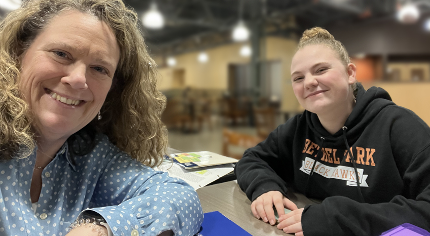 Two people sitting at a table in a large indoor space. The person on the left has curly hair and wears a blue polka dot shirt. The person on the right has tied-up hair and is wearing a black hoodie with orange lettering. They are both smiling.