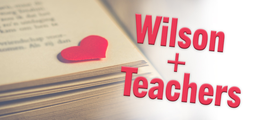 A stack of papers with a small red heart on top. The text Wilson + Teachers is in bold red letters on the right. The background is softly blurred, highlighting the text and heart.