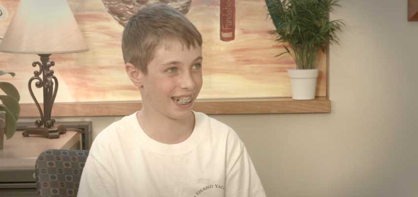 A smiling boy with braces sitting in a room, wearing a white T-shirt. A framed painting, a potted plant, and a lamp are visible in the background.