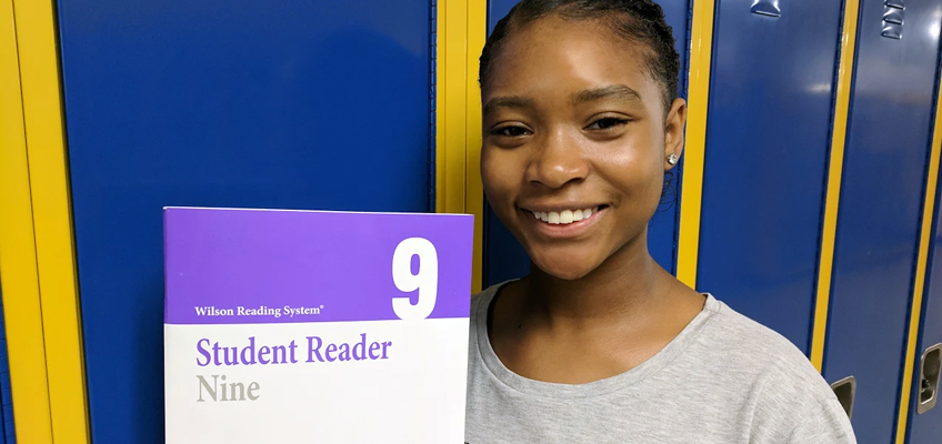 A person smiling while holding up a book titled Student Reader Nine from the Wilson Reading System. They are standing in front of a row of blue and yellow lockers.