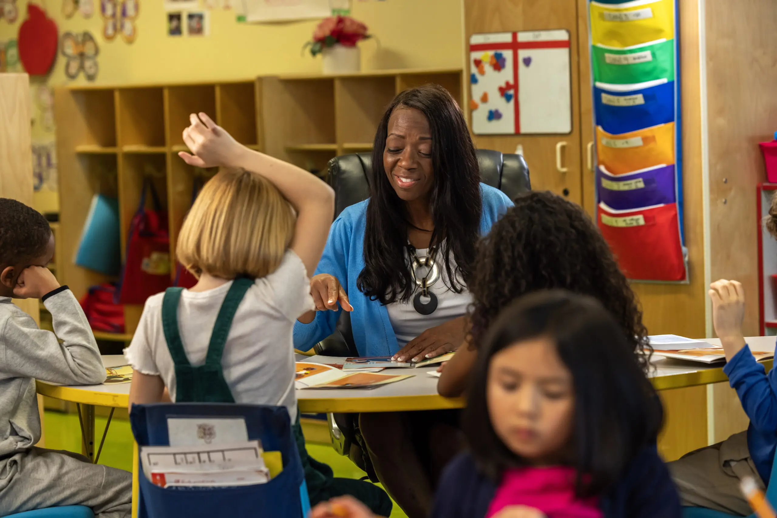 A teacher sits at a round table with young students in a classroom. One child raises their hand, while others engage with books or papers. Colorful decorations and storage are visible in the background.