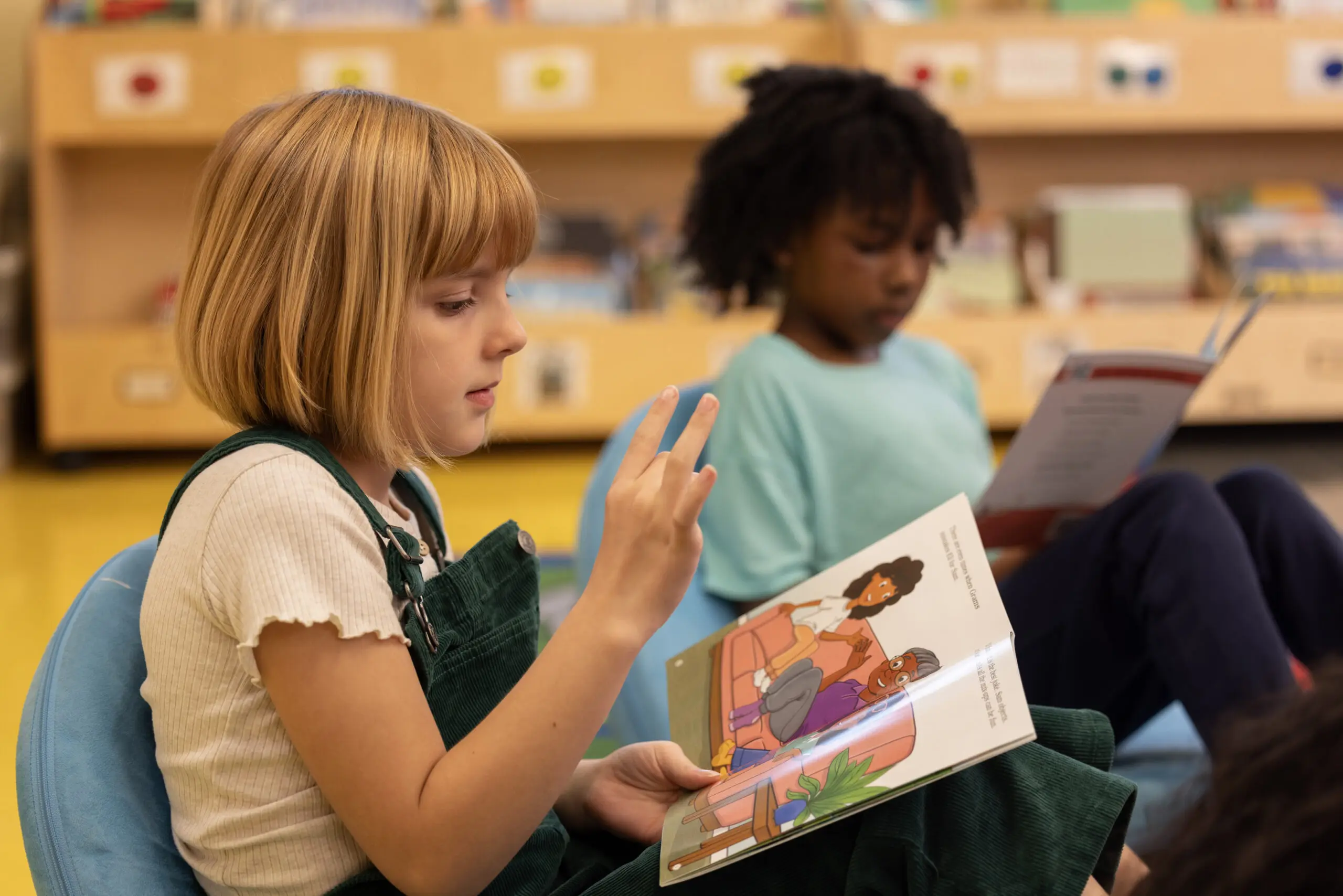 Two children are sitting on the floor, reading picture books in a classroom. The child in the foreground, with short blonde hair, is wearing green overalls and gesturing with their hand. The background is slightly blurred, showing bookshelves.