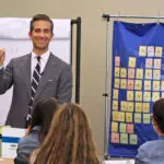 A man in a suit is standing in front of a group, holding a pen. Behind him is a flip chart and a blue chart displaying letters and words. Several people, seen from the back, are seated and watching him.