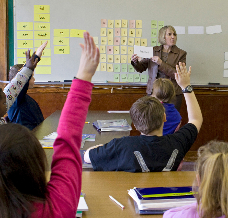 Teacher holding up a word card in front of a classroom and students are raising their hands