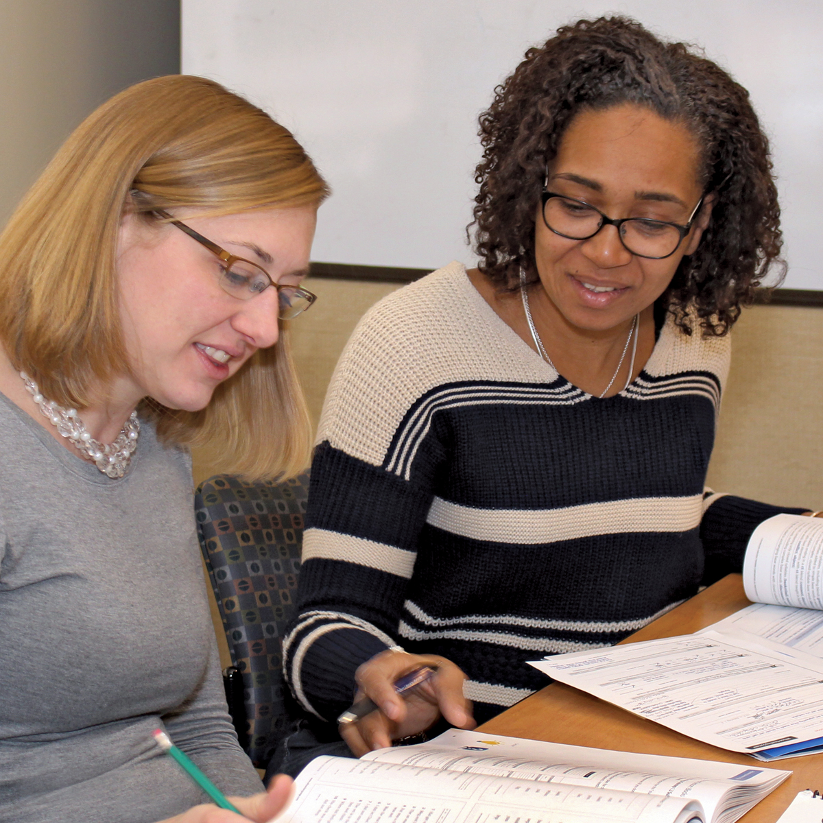 Woman with dark hair explaining something from a book to a woman with blonde hair. 