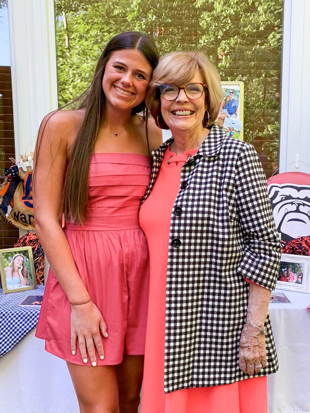 A teenage girl stands in a pink dress stands next to a senior woman in a  pink dress and black and white checked jacket. Both people are smiling.