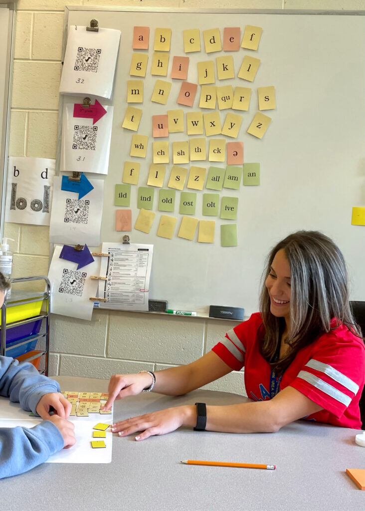 Female teacher with long brown hair and a red Tshirt sitting at a table explaining something to a child who is out of the frame.