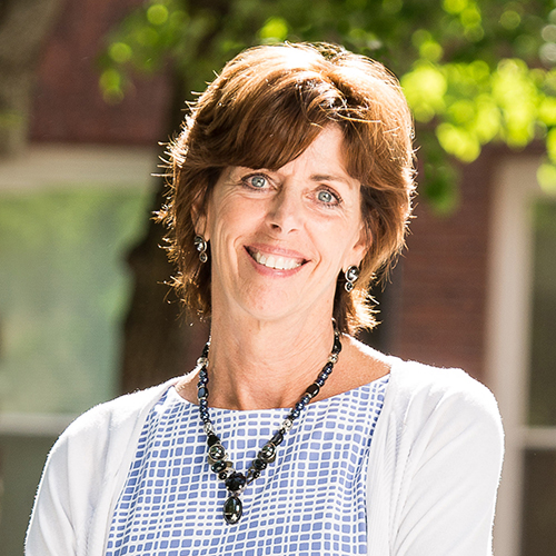 Smiling woman with short brown hair stands outdoors wearing a black necklace, earrings, blue and white checkered top, and a white cardigan. The background is blurred with greenery and a brick wall visible.