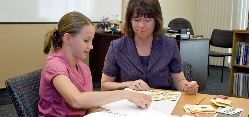 A young girl in a pink shirt works on an educational activity with an older woman in a purple outfit. They are seated at a table in an office, using cards and papers, with shelves and chairs in the background.