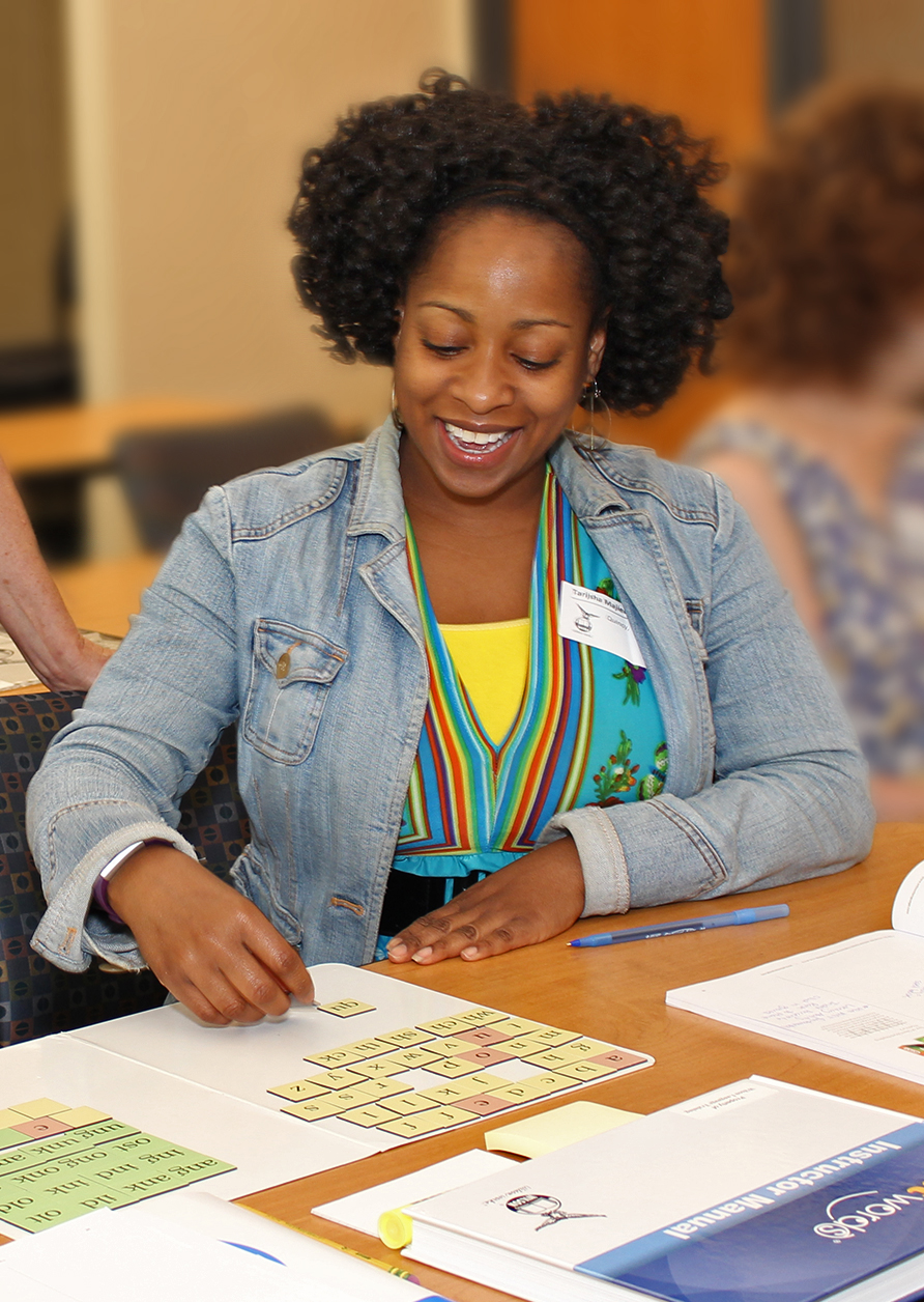 A woman with curly hair, wearing a denim jacket and a colorful striped shirt, smiles while engaging in an educational activity at a table. Various papers, a pen, and a book are spread out in front of her. Another person is partially visible in the background.