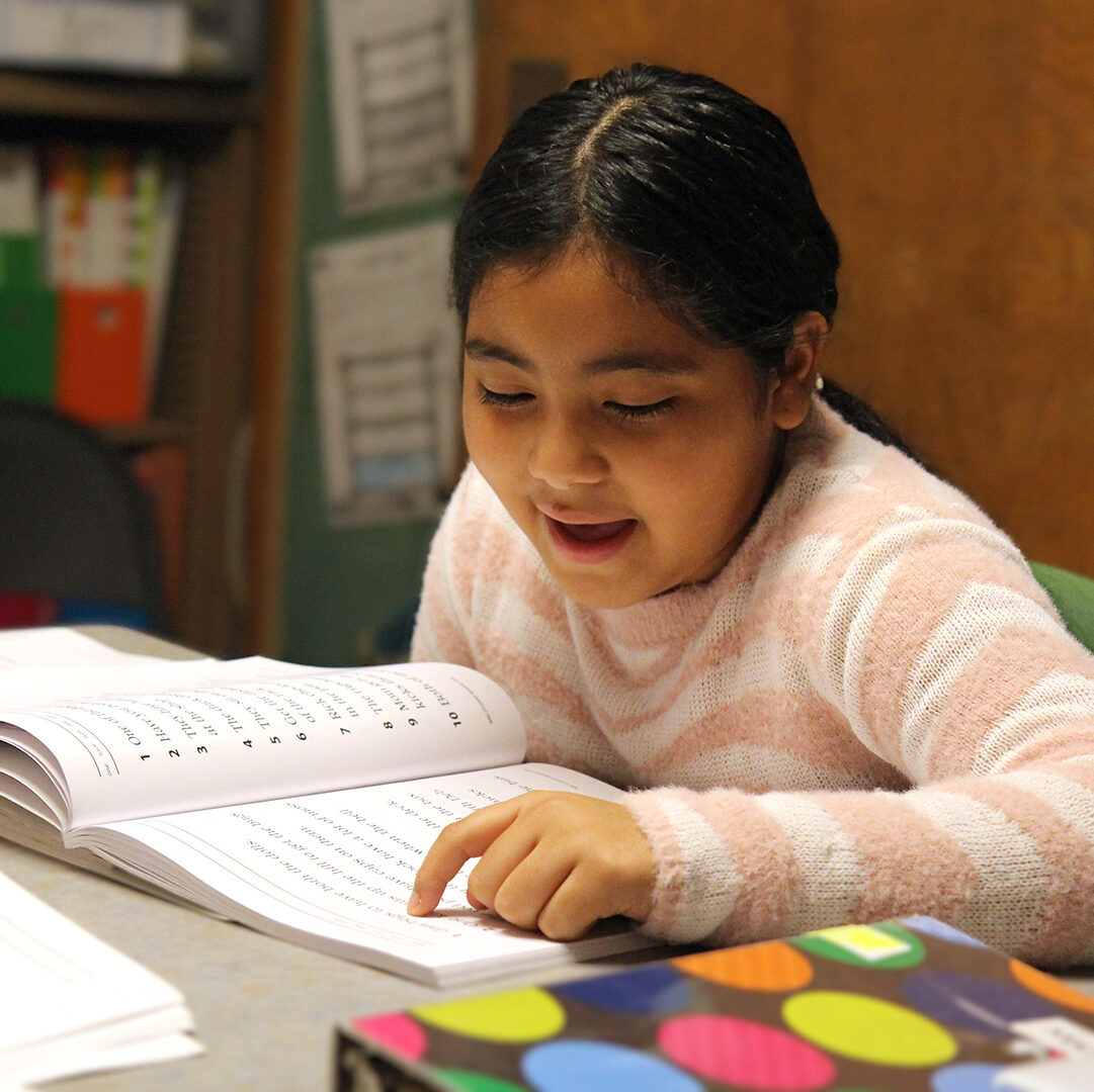 A young girl with dark hair is sitting at a table, reading aloud from an open textbook. She points to the text with her finger. An open laptop and colorful binders are in the background. Another person’s hand holding a pen is visible on the left side of the image.
