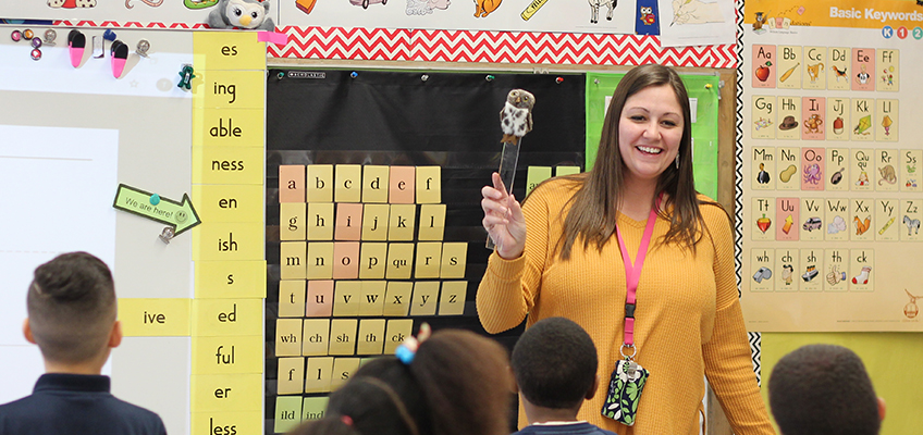 A teacher in a yellow sweater holds a pointer and smiles while standing in front of a classroom. Children sit facing her. The background shows educational posters with letters and words on the walls.