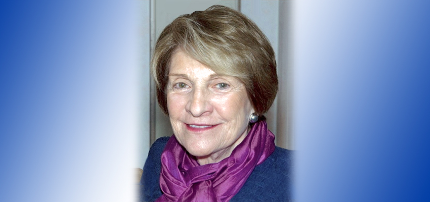 An older woman with short light brown hair is smiling. She is wearing a dark blue outfit and a purple scarf. The background has a gradient from white to blue on the sides.