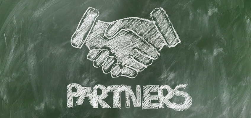Chalk drawing of two hands shaking above the word PARTNERS on a green chalkboard background.