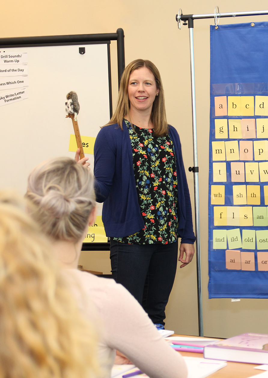 A woman wearing a floral shirt stands at the front of a classroom, holding a small stuffed animal on a stick. She faces students who are seated. Behind her is a board with the alphabet and word endings on colorful cards.