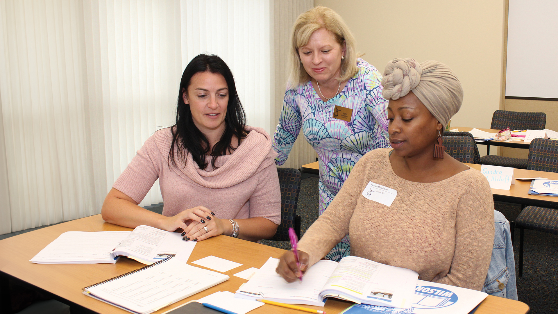 Three women are gathered at a table. Two are seated, taking notes from open books, while the third stands over them, observing. They appear to be in a classroom or meeting setting. One woman wears a brown head wrap and a name tag.