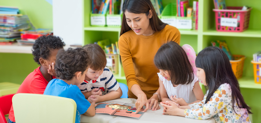 A teacher is sitting at a table with five young children, reading a colorful book together. The background shows shelves filled with books in a classroom. The children are engaged and listening attentively.