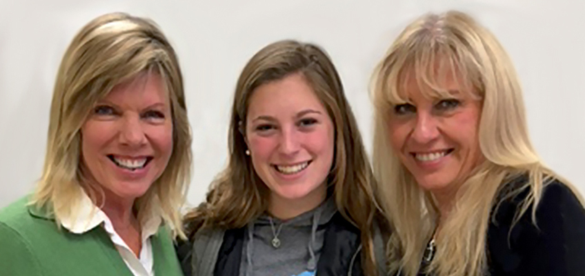 Three women are smiling at the camera. The woman on the left has short blonde hair and wears a green sweater. The woman in the center has long brown hair and wears a gray top. The woman on the right has long blonde hair and wears black.