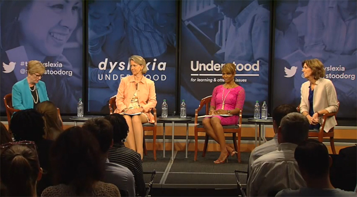 Four women seated on stage for a panel discussion about dyslexia. They are surrounded by banners with text related to dyslexia and learning. An audience is visible in the foreground.