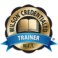 The image is a badge with the words "Wilson Credentialed Trainer" encircling an open book symbol. A blue ribbon below reads "Trainer" and "W.C.T." is at the bottom of the badge.