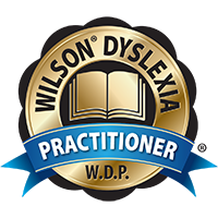A gold and blue circular badge featuring an open book in the center, surrounded by the text "Wilson Dyslexia Practitioner W.D.P." in uppercase letters. A ribbon at the bottom of the badge displays the word "Practitioner" prominently.