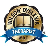A gold and black emblem with the text "Wilson® Dyslexia" around the top, an open book illustration in the center, and a blue ribbon with the word "Therapist" in white letters across the bottom. The initials "W.D.T." appear at the bottom of the emblem.