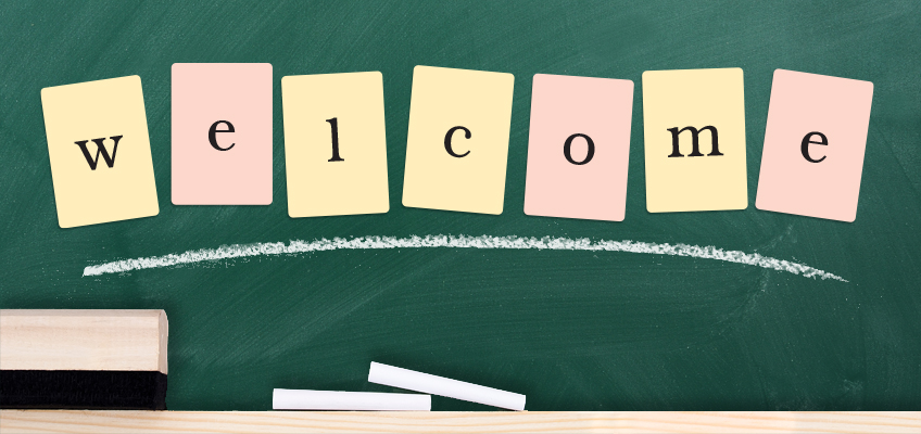 The image shows letters on pastel-colored cards spelling welcome against a green chalkboard. Below the letters is a white chalk line, with three pieces of white chalk resting on a wooden chalkholder.