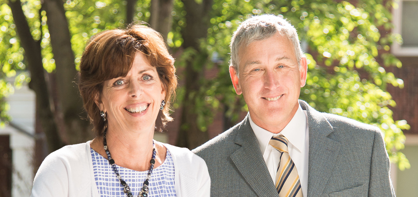 A woman and a man are smiling at the camera outdoors. The woman is wearing a white and blue patterned top with a necklace, and the man is in a gray suit with a striped tie. Green trees are visible in the sunny background.