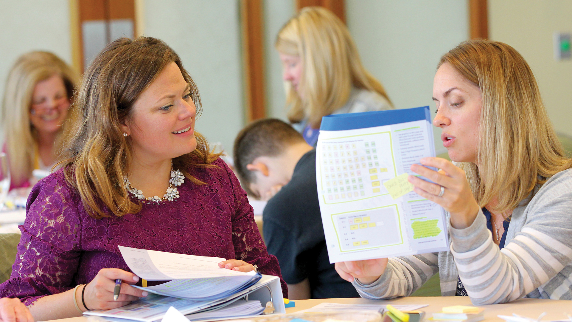 Two women sit at a table, engaged in conversation and looking at a document with highlighted sections. They are surrounded by others at a workshop or meeting. The atmosphere is collaborative and focused.