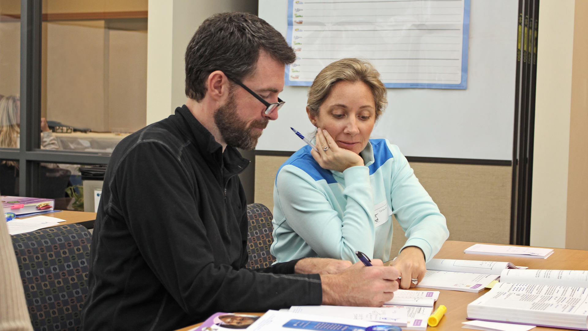 A man and a woman sit at a table in an office, reviewing documents together. The man is writing with a pen, while the woman attentively observes, resting her chin on her hand. Papers and a highlighter are spread out on the table.