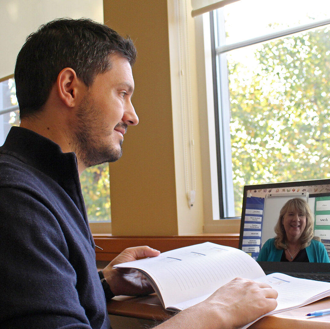 A man sits by a window, holding an open book, and looks at a woman on a laptop screen. The laptop shows a video call with the woman smiling. Sunlight filters through the window, illuminating the scene.