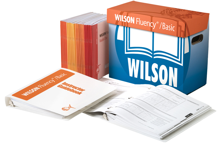 A Wilson Fluency/Basic kit is displayed, including a large box, a set of teacher's books, and an open instructor guidebook. The box and materials are branded with the Wilson logo and designed for educational purposes.