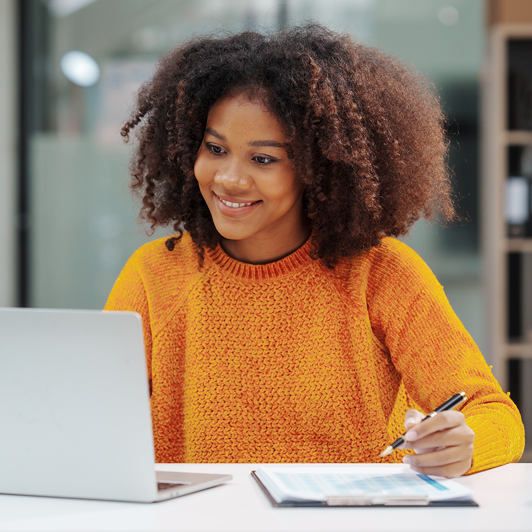 A person with curly hair, wearing an orange sweater, is smiling while looking at a laptop. They are holding a pen and a clipboard with a document, sitting at a white table in an indoor setting.