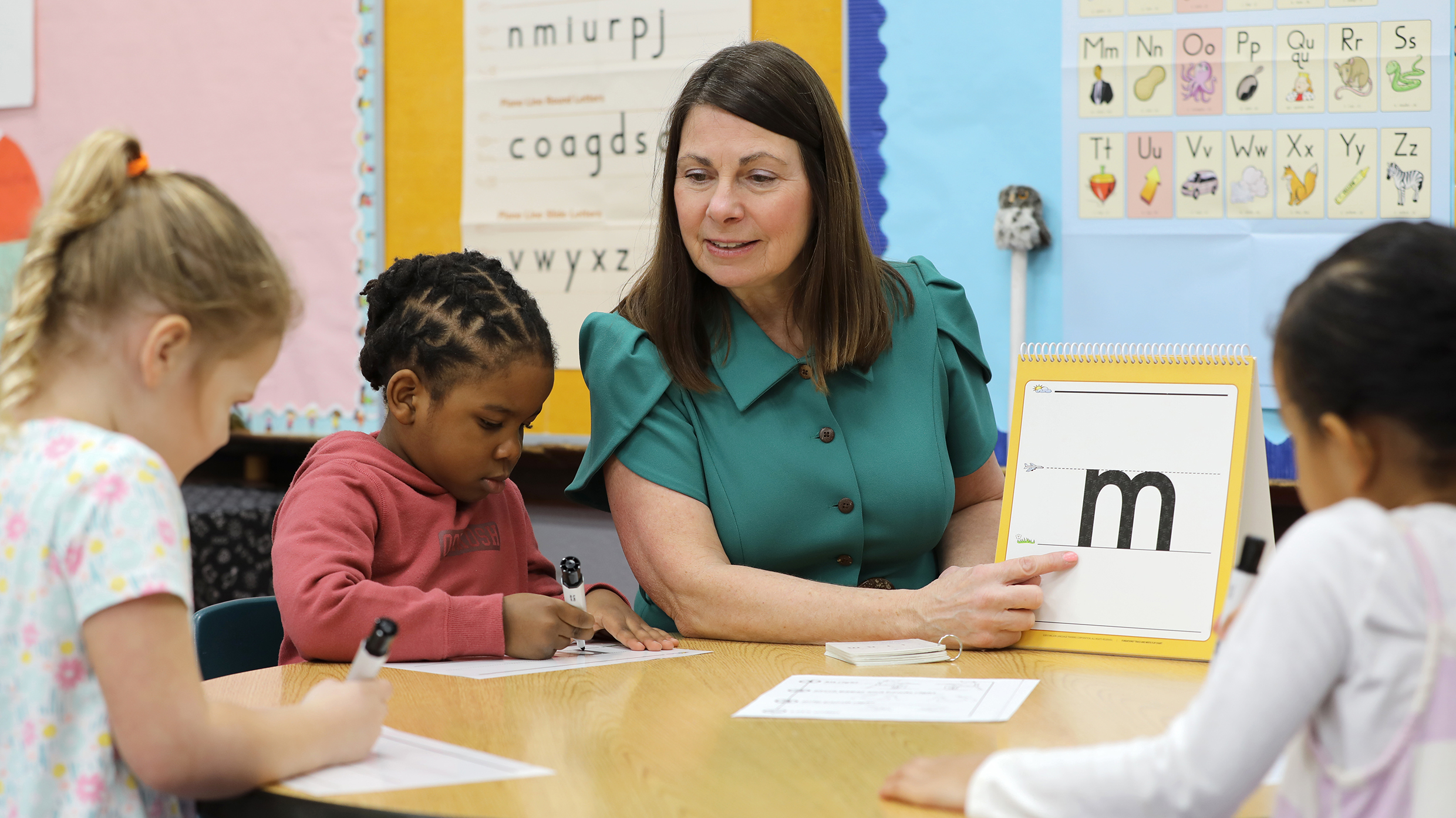 A teacher sits at a round table showing a flashcard with the letter m to three young students who are working on their own tasks. The classroom is decorated with educational posters, including an alphabet chart on the wall.