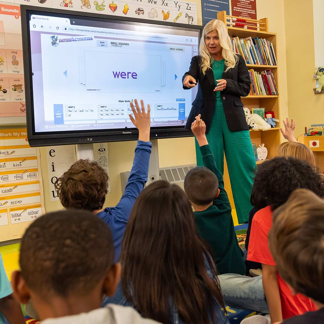 A teacher stands in front of a large interactive screen displaying the word were with several images and words. She is pointing at the screen while several young students seated on the floor raise their hands. Bookshelves and educational posters decorate the classroom.